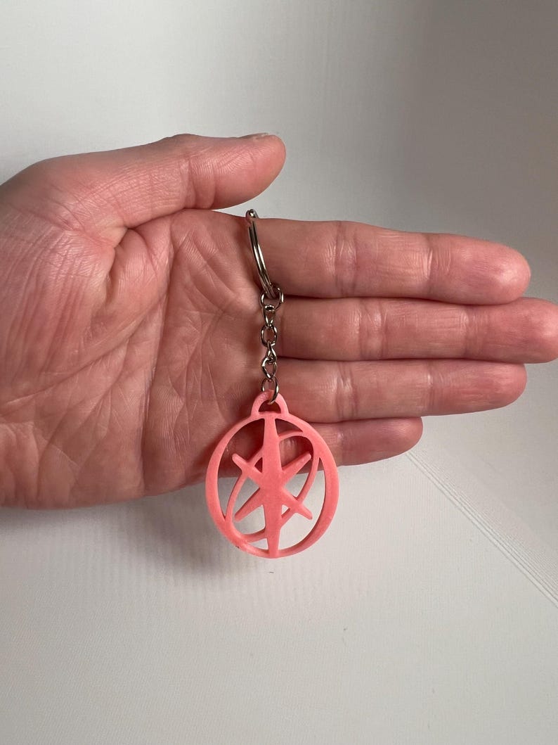 May include: A light pink keychain with a circular design and a starburst-like pattern inside. The keychain is attached to a silver chain and ring. The design is a simple, geometric shape.