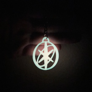 May include: A glowing, circular pendant with a starburst design. The pendant is a light, bright blue color and is suspended from a small chain. The background is dark, emphasizing the pendant's luminescence.