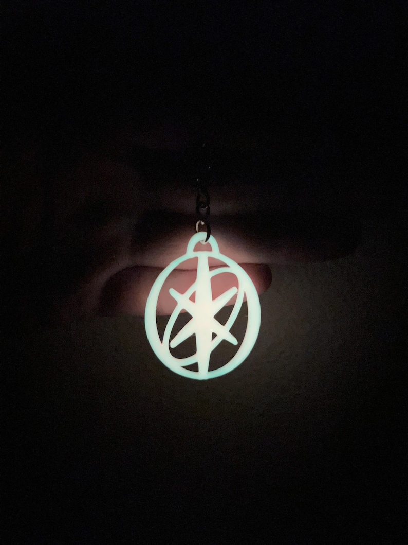May include: A glowing, circular pendant with a starburst design. The pendant is a light, bright blue color and is suspended from a small chain. The background is dark, emphasizing the pendant's luminescence.