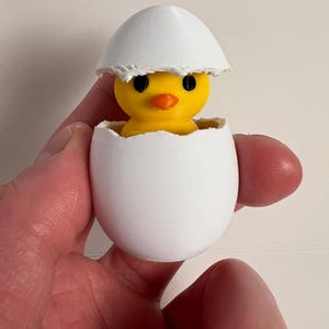 May include: A yellow chick figurine with a bright orange beak and black eyes peeks out of a white eggshell. The top of the eggshell is open, revealing the chick. The figurine is held in a hand.