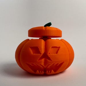 May include: An orange 3D-printed pumpkin with a jack-o'-lantern face. The pumpkin has a removable lid and a small green stem. The design features carved-out eyes, a nose, and a mouth, perfect for Halloween decorations.