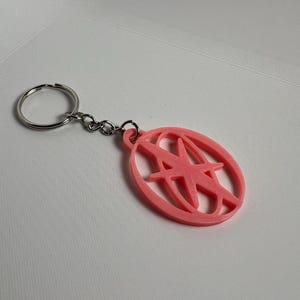 May include: A pink keychain with a silver key ring and chain. The keychain features a pink oval with a stylized letter "A" design inside. The keychain is made of a solid material and is likely 3D printed.