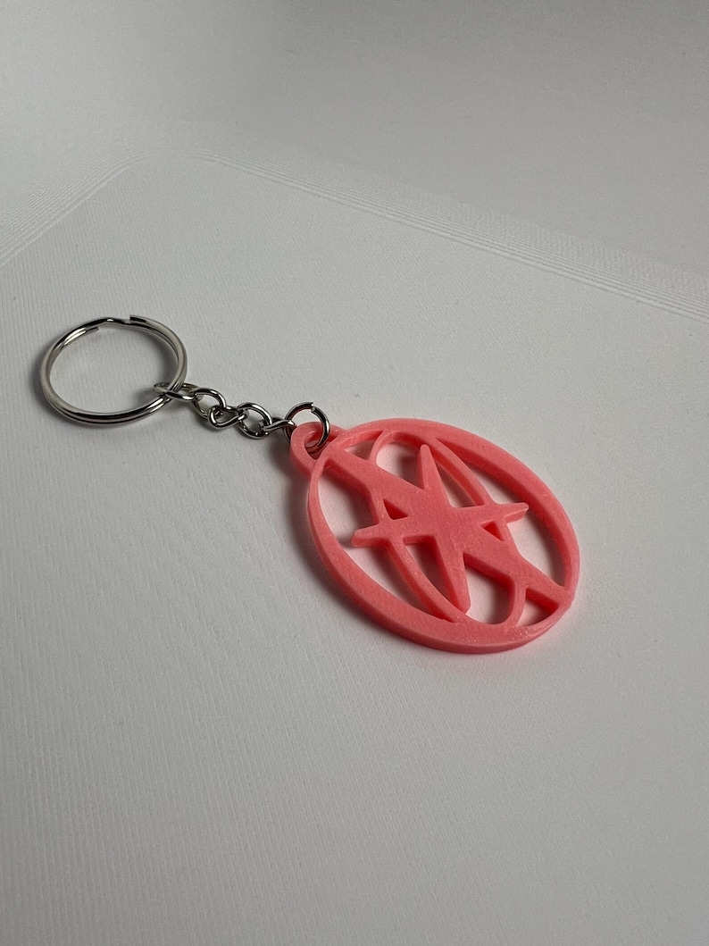 May include: A pink keychain with a silver key ring and chain. The keychain features a pink oval with a stylized letter "A" design inside. The keychain is made of a solid material and is likely 3D printed.