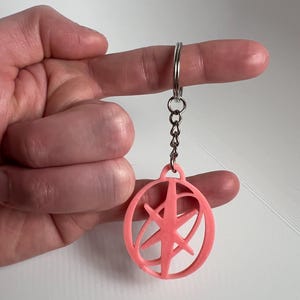 May include: A pink keychain with a circular design and a starburst pattern inside. The keychain is attached to a silver chain and ring. The design is a solid, three-dimensional shape.