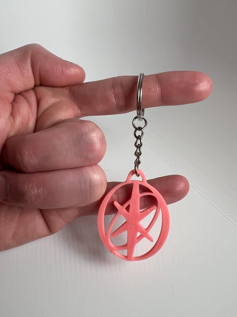 May include: A pink keychain with a circular design and a starburst pattern inside. The keychain is attached to a silver chain and ring. The design is a solid, three-dimensional shape.