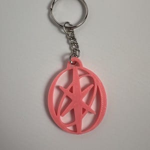 May include: A pink keychain with a circular pendant featuring a starburst design. The pendant is attached to a silver chain and keyring. The keychain is made of plastic and is likely 3D printed.