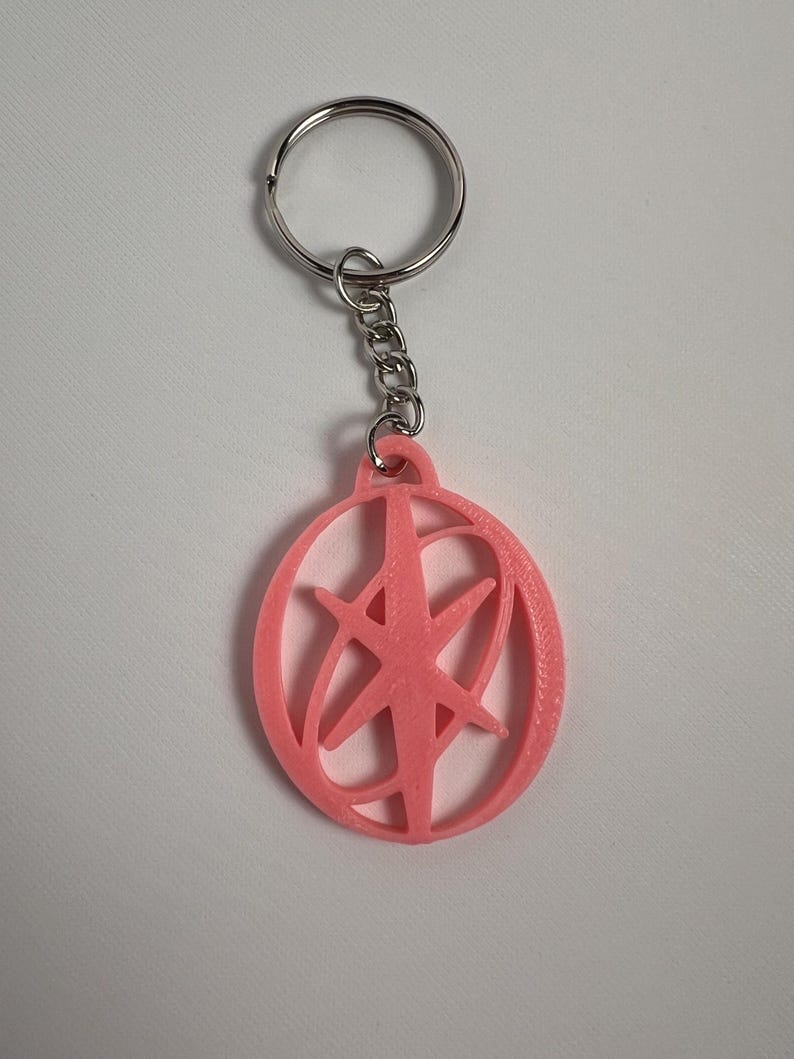 May include: A pink keychain with a circular pendant featuring a starburst design. The pendant is attached to a silver chain and keyring. The keychain is made of plastic and is likely 3D printed.