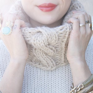 Pode incluir: Um close-up de uma mulher vestindo um suéter de tricô de cabo bege com gola alta. Ela está usando vários anéis de ouro nos dedos e uma pilha de pulseiras de ouro no pulso.