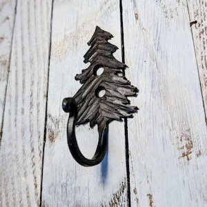 May include: A black metal wall hook shaped like a stylized Christmas tree. The hook has a circular loop and is mounted on a distressed white wooden surface. The tree design features textured details and two circular cutouts.