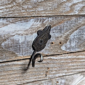 May include: A decorative, dark metal hook with a leaf-shaped top and a curved hook. The hook is attached to a wooden surface with a screw. The wood has a weathered appearance with visible grain and white paint.