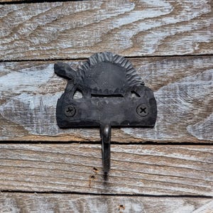 May include: A black metal wall hook with a decorative sunburst design. The hook is attached with two screws and has a single hook for hanging items. The hook is mounted on a weathered wooden surface.