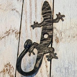 May include: A dark gray metal gecko-shaped hook. The hook has a textured surface and a circular hole near the center. The gecko's tail curves into a hook shape. The gecko's feet and head are detailed. The hook is mounted on a light wood surface.
