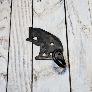 May include: A black metal cat-shaped wall hook. The hook is in the shape of a cat grooming itself. The cat has two holes and a curled tail. The hook is mounted on a white wooden surface.