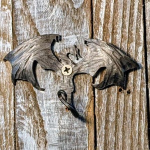 Puede incluir: Un gancho de pared de metal con forma de dragón gris oscuro, con alas extendidas y un gancho curvado, fijado a una superficie de madera con un tornillo. La madera tiene un aspecto envejecido. El dragón es detallado.