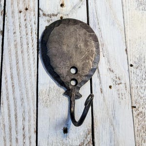 May include: Hand-forged metal balloon-shaped wall hook. The hook is dark gray with two circular holes near the top and a curved hook at the bottom. The metal has a textured surface and is mounted on a white wooden background.