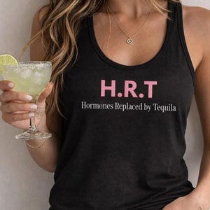 以下が含まれることがあります： 前面にピンク色の「H.R.T」という文字と、白色の「Hormones Replaced by Tequila」という文字がプリントされた黒色のレーサーバックタンクトップ。青色のデニムジーンズと、小さな円形のペンダントが付いた繊細なゴールドのネックレスを合わせて着用しています。
