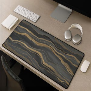 Pode incluir: Um tapete de secretária retangular com um fundo cinzento-carvão escuro e linhas diagonais abstratas e fluidas em dourado cintilante e cinzento claro. O tapete tem um rebordo preto cosido e está colocado sobre uma superfície de madeira.