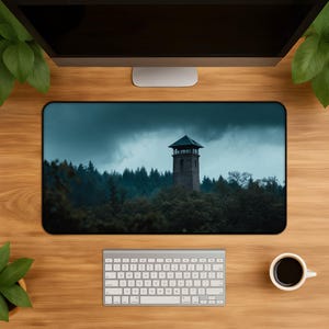 May include: A rectangular desk mat featuring a high-resolution print of a stone watchtower rising above a dense, dark forest under a moody, overcast sky. The mat is positioned on a wooden desk surface, centered below a computer monitor and keyboard.