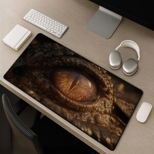May include: A rectangular desk mat featuring a high-resolution, close-up graphic of a reptilian eye with a vertical pupil. The iris is a vibrant amber-orange colour, surrounded by dark, textured, scaly skin in shades of brown and black.
