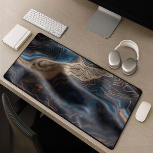 May include: A rectangular desk mat featuring an abstract topographic map design with flowing lines in shades of deep blue, dark charcoal, and warm tan. The mat has a smooth surface and black stitched edges, positioned on a light wood grain desk.