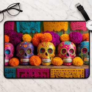 May include: A rectangular mousepad featuring colorful Day of the Dead sugar skulls. The skulls are painted in vibrant hues of pink, yellow, purple, and blue, with intricate floral designs. Orange and purple pom-poms and decorative cut paper complete the festive scene.