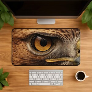 Op de afbeelding: Een rechthoekige bureaumat met een hoge resolutie close-up print van het oog van een roofvogel. Het ontwerp toont gedetailleerde bruine en beige veren rondom een scherpe, goudgele iris met een zwarte pupil en een gebogen gele snavel.