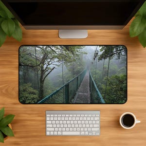 May include: A rectangular desk mat featuring a high-resolution print of a narrow metal suspension bridge stretching through a dense, misty green forest. The scene depicts a walkway with green railings surrounded by lush trees and thick fog.