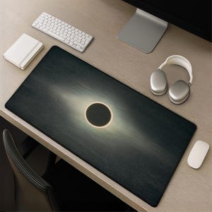Puede incluir: Alfombrilla de escritorio rectangular negra con un diseño celestial, con un círculo central oscuro rodeado por un anillo más claro y un fondo nublado. También se ven un teclado, un ratón y unos auriculares blancos.