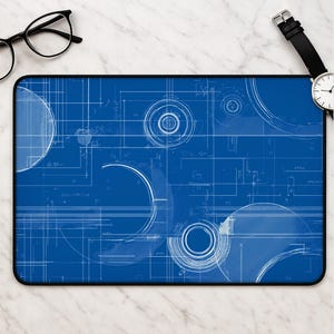 Puede incluir: Una alfombrilla de escritorio rectangular con un fondo azul intenso y líneas de plano técnico blancas, formas geométricas y círculos concéntricos. El diseño incluye varias líneas que se cruzan y patrones de dibujo técnico en la superficie.