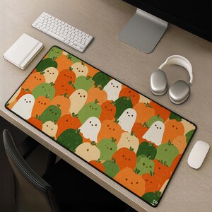 Op de afbeelding: Een rechthoekige bureaumat met een herhalend patroon van kleine, cartoonachtige spoken en pompoenen in tinten oranje, groen en crème. De spoken hebben stipogen en kleine glimlachjes, terwijl de pompoenen groene steeltjes hebben.