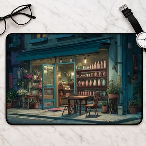 May include: A rectangular desk mat featuring an illustration of a storefront at dusk with a teal awning, a wooden table and chairs, shelves filled with ceramic vases, and potted plants, positioned next to black-rimmed glasses and a wristwatch.