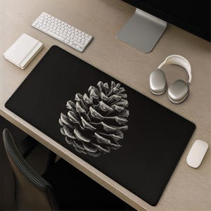 May include: A rectangular black desk mat featuring a large, detailed white line-art illustration of a pinecone centered on the surface. The mat has a smooth, dark fabric texture with a clean, stitched edge, providing a stark contrast to the white botanical design.