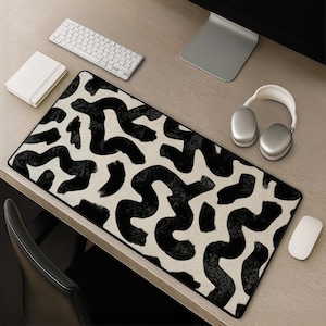 May include: A rectangular desk mat featuring a repeating pattern of thick, black, hand-painted style wavy lines and abstract shapes set against a solid off-white background. The mat has a black stitched border and a smooth, flat surface texture.