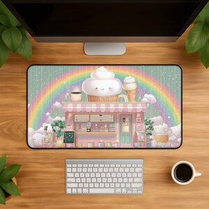 May include: A rectangular desk mat featuring a pastel pink cafe shop with a large smiling cupcake on the roof, a vibrant rainbow arching across the background, scattered clouds, and a menu board, all set against a soft, whimsical, light-colored sky.