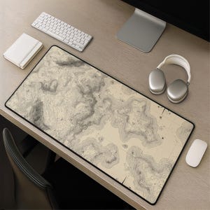 Puede incluir: Una alfombrilla de escritorio rectangular con un diseño de mapa topográfico detallado en tonos beige, crema y gris oscuro. La superficie muestra líneas de contorno intrincadas que representan cambios de elevación, con un borde negro.