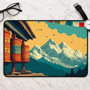 May include: A rectangular desk mat featuring a vibrant illustration of orange prayer wheels in the foreground, set against a backdrop of snow-capped mountain peaks under a yellow sky, with colorful prayer flags strung across the upper portion.