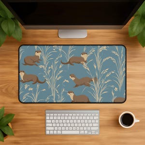 May include: A rectangular mouse pad featuring a repeating pattern of brown otters and beige grass-like plants on a blue background. The mouse pad is rectangular with a black border. The image is set on a wooden desk with a keyboard and coffee cup.