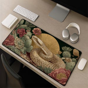 May include: A rectangular desk mat featuring a detailed illustration of a coiled snake in the centre, surrounded by green cacti and large red flowers. A golden sunburst design sits behind the snake against a dark background, creating a contrast.