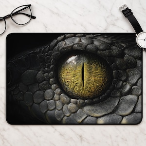 May include: A rectangular desk mat featuring a high-resolution, close-up graphic of a reptile eye with a vertical slit pupil. The iris is a textured, golden-yellow colour, surrounded by dark, overlapping scales in shades of black and charcoal grey.