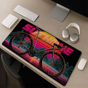 Puede incluir: Una alfombrilla de escritorio rectangular con fondo negro y un gráfico de colores neón de una bicicleta de carretera. El diseño incluye un gran sol amarillo, rayas horizontales rosas y montañas de estructura alámbrica azul con el texto SUTHOKE en rosa.