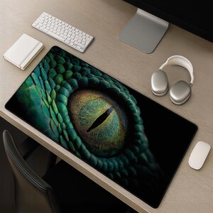 Könnte beinhalten: Eine rechteckige Schreibtischunterlage mit einem hochauflösenden Grafikdruck eines Reptilienauges in Nahaufnahme. Das Design zeigt eine zentrale vertikale Pupille, umgeben von einer texturierten, schillernden grün-goldenen Iris und dunklen Schuppen.