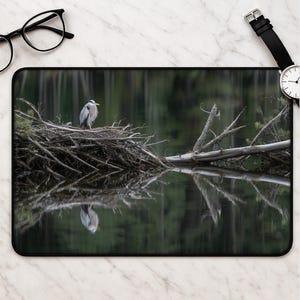 May include: A rectangular desk mat featuring a high-resolution photograph of a grey heron perched on a large, tangled nest of branches situated in a dark, still body of water, with the bird's reflection clearly visible on the water's surface.