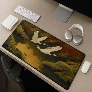 Op de afbeelding: Een rechthoekige bureaumat met een gedetailleerde illustratie van twee witte kraanvogels in vlucht boven een donker, reflecterend wateroppervlak, omringd door dicht, getextureerd groen en donkerrood gebladerte met een zwarte rand.