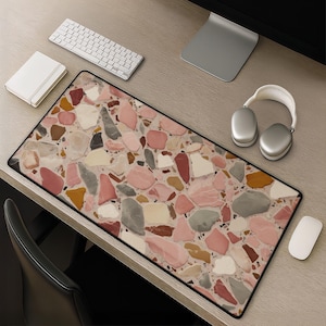 May include: A rectangular desk mat with a terrazzo pattern in shades of pink, gray, and beige. The mat is surrounded by a black border. A white keyboard, mouse, headphones, and notepad are on the desk.
