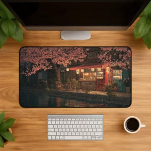May include: A rectangular desk mat featuring a detailed illustration of a Japanese street scene at night, with a canal, a small shop with red awnings, glowing lanterns, and cherry blossom trees with pink flowers hanging over the water.