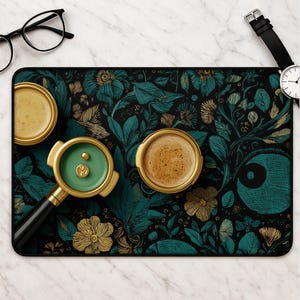 May include: A rectangular desk mat featuring a dark background with teal and gold botanical illustrations. The mat holds two round cups of coffee and a gold-toned espresso portafilter with a green interior, alongside black-rimmed glasses.