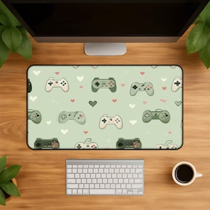 May include: A rectangular desk mat featuring a light sage green background with a repeating pattern of various game controllers and small white and pink hearts. The design includes multiple controller styles in shades of green and white.
