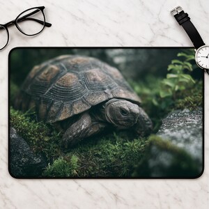 May include: A rectangular desk mat featuring a high-resolution photograph of a brown tortoise walking through green moss and grey rocks. The mat has a black border and is displayed alongside a pair of black-rimmed glasses and a wristwatch.