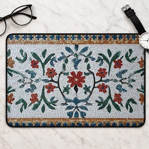 May include: A rectangular desk mat featuring a detailed mosaic pattern of red flowers, green leaves, and blue accents on a white background, framed by a border of tan and blue tiles, accompanied by black-rimmed glasses and a black wristwatch.