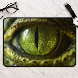 Peut inclure: Un tapis de souris rectangulaire présentant un graphique haute résolution et détaillé d'un œil de reptile avec une pupille noire verticale et des écailles texturées vert olive entourant l'iris, sur un fond à motif marbre blanc.